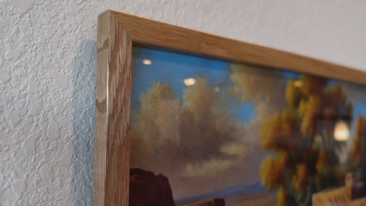 Load video: A video showcasing the Whispers of the Southwest painting in two different frames and sizes. The larger 24x36-inch black frame leans against a counter, while the smaller 12x18-inch oak frame is hung above it. The camera captures various angles of the artwork, highlighting the textures and details. A potted plant sits to the left of the black frame, and a lamp is positioned to its right, creating a warm and balanced setting.